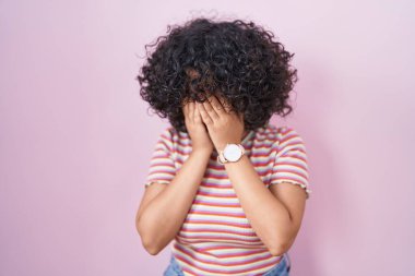 Orta Doğulu genç bir kadın pembe arka planda duruyor. Ağlarken suratını ellerle kapatıyor. depresyon kavramı. 