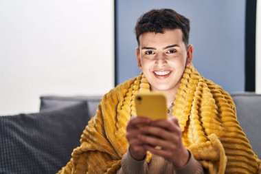Young non binary man using smartphone sitting on sofa at home