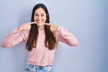 Genç esmer kadın mavi arka planda durup neşeli bir şekilde gülümseyerek parmaklarını ve ağzını işaret ediyor. diş sağlığı kavramı. 