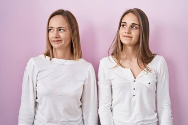 Middle age mother and young daughter standing over pink background smiling looking to the side and staring away thinking. 