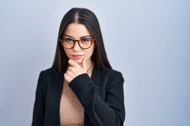 Young brunette woman standing over blue background looking confident at the camera with smile with crossed arms and hand raised on chin. thinking positive. 