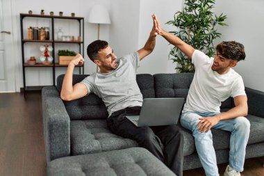 Two hispanic men couple using laptop high five with hands raised up at home