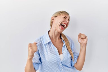 Güzel sarışın bir kadın beyaz arka planda mutlu ve heyecanlı bir şekilde kollarını kaldırıp, gülümseyerek ve başarı için bağırarak zafer jesti yapıyor. kutlama konsepti. 