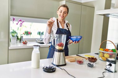 Genç sarışın kadın mutfaktaki blendere buz dökerek gülümsüyor.