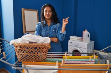 Young asian woman hanging clothes at clothesline with a big smile on face, pointing with hand and finger to the side looking at the camera. 