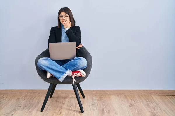 Young hispanic woman sitting on chair using computer laptop looking stressed and nervous with hands on mouth biting nails. anxiety problem.