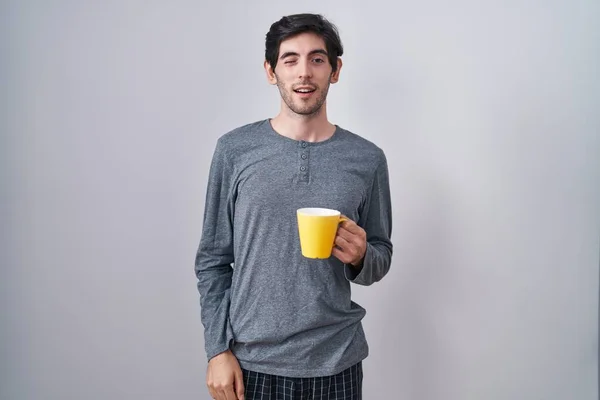 Young hispanic man wearing pajama drinking a cup of coffee winking looking at the camera with sexy expression, cheerful and happy face.