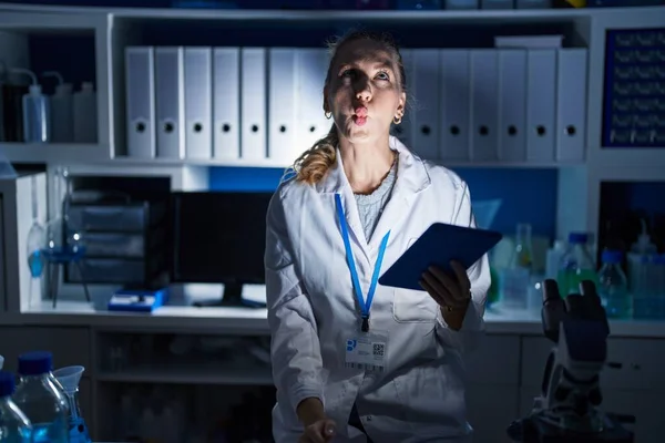 Beautiful blonde woman working at scientist laboratory late at night making fish face with lips, crazy and comical gesture. funny expression.
