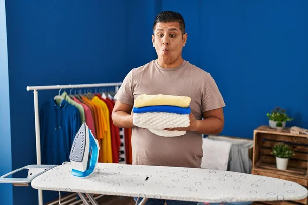 Hispanic young man holding folded laundry after ironing making fish face with mouth and squinting eyes, crazy and comical.