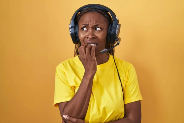 African american woman listening to music using headphones looking stressed and nervous with hands on mouth biting nails. anxiety problem.