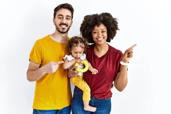 Interracial young family of black mother and hispanic father with daughter cheerful with a smile on face pointing with hand and finger up to the side with happy and natural expression