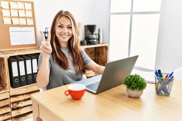 在办公室里工作的年轻的黑发女人 拿着笔记本电脑 用手指指了指二号 带着自信和快乐的笑容 — 图库照片
