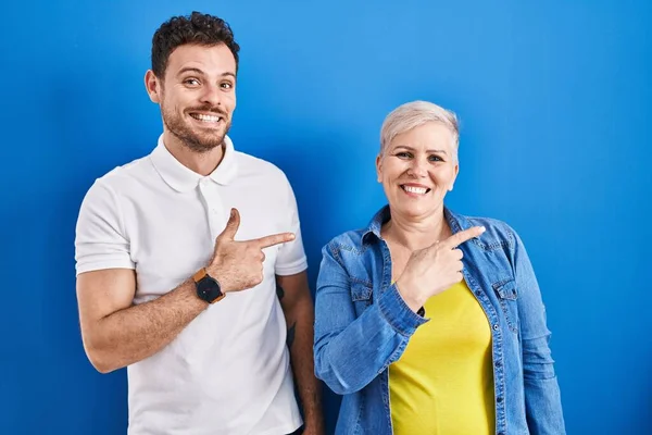 若いブラジル人の母親と息子は 幸せと自然な表情で側に手と指を指して笑顔で陽気に青い背景に立っています — ストック写真