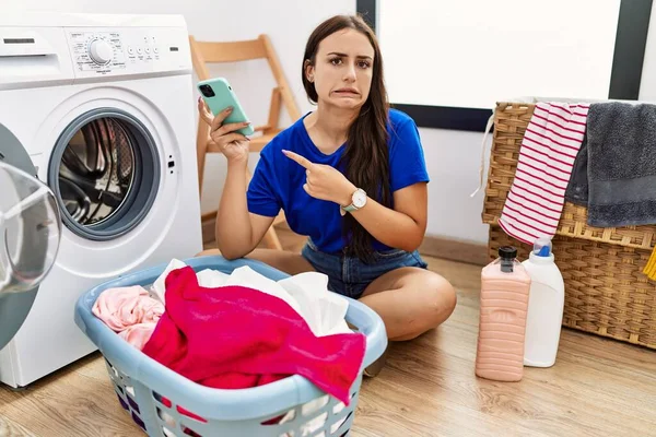 若いですブルネット女性を使用して洗濯スマートフォンを指して脇に心配と神経で前指 懸念と驚きの表現 — ストック写真
