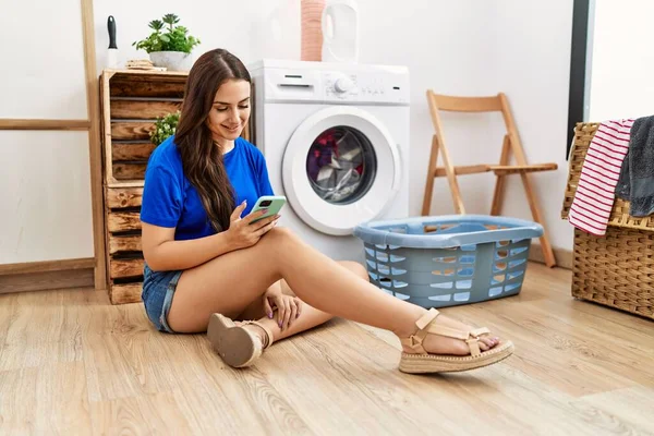 洗濯室で洗濯機を待っているスマートフォンを使っている若い白人女性 — ストック写真