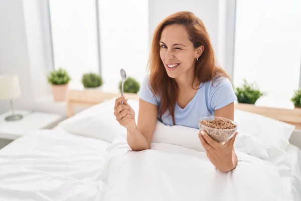 Brunette woman eating breakfast in the bed winking looking at the camera with sexy expression, cheerful and happy face.