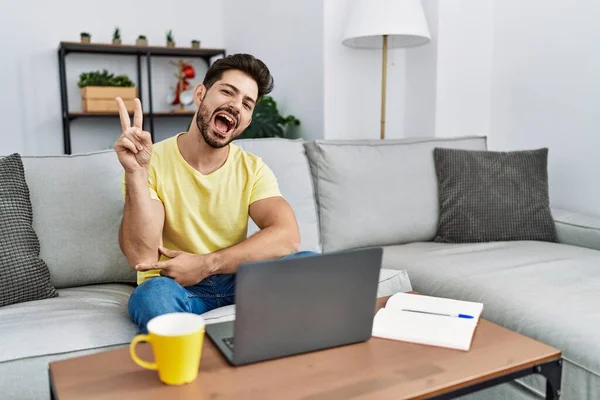 Young man with beard using laptop at home smiling with happy face winking at the camera doing victory sign. number two.