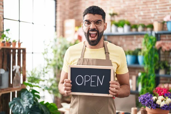 Hispanic man with beard working at florist holding open sign angry and mad screaming frustrated and furious, shouting with anger. rage and aggressive concept.