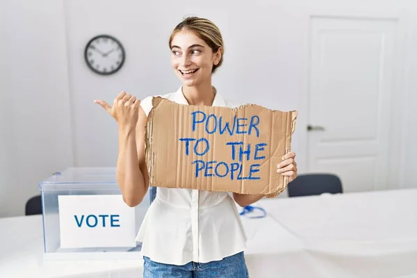 Young blonde woman at political election holding power to the people banner pointing thumb up to the side smiling happy with open mouth