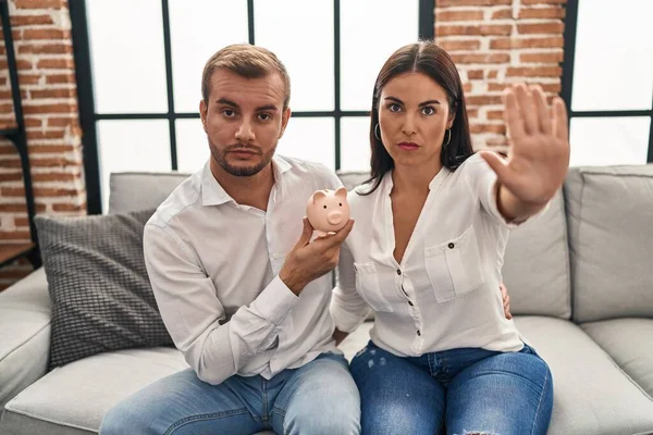 Young hispanic couple holding piggy bank sitting on the sofa with open hand doing stop sign with serious and confident expression, defense gesture