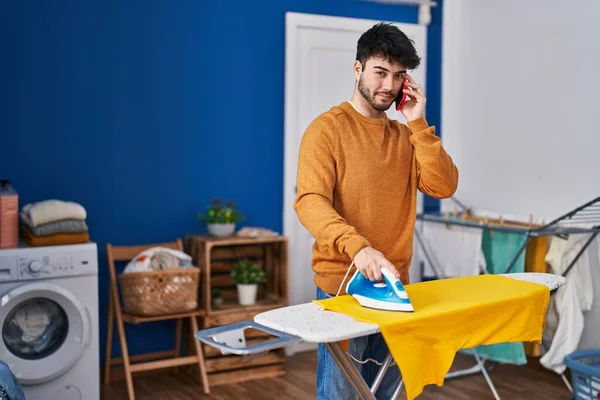 Young Hispanic Man Talking Smartphone Ironing Clothes Laundry Room — Φωτογραφία Αρχείου