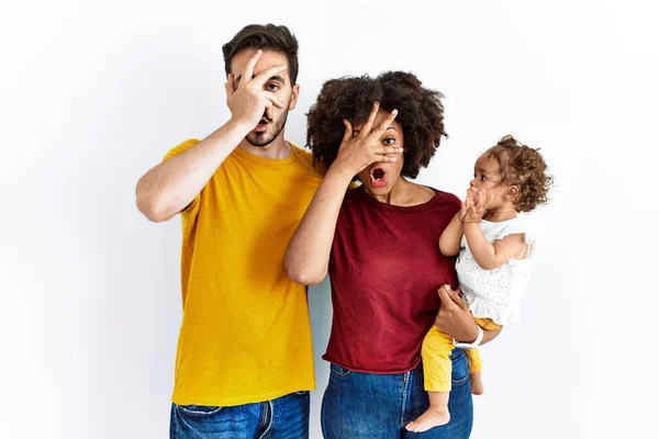 Interracial young family of black mother and hispanic father with daughter peeking in shock covering face and eyes with hand, looking through fingers with embarrassed expression.