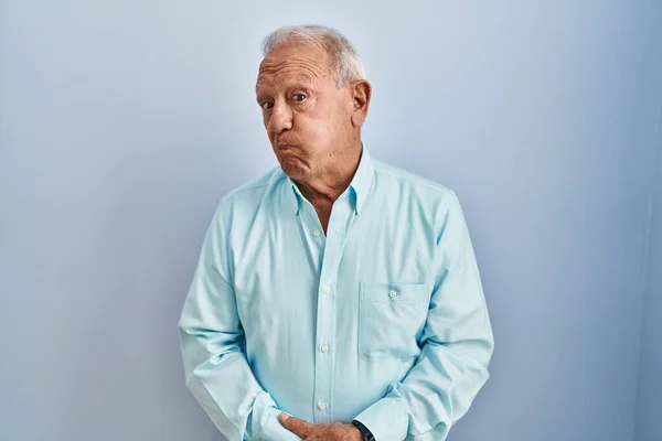Homem Sênior Com Cabelos Grisalhos Sobre Fundo Azul Soprando Bochechas — Fotografia de Stock