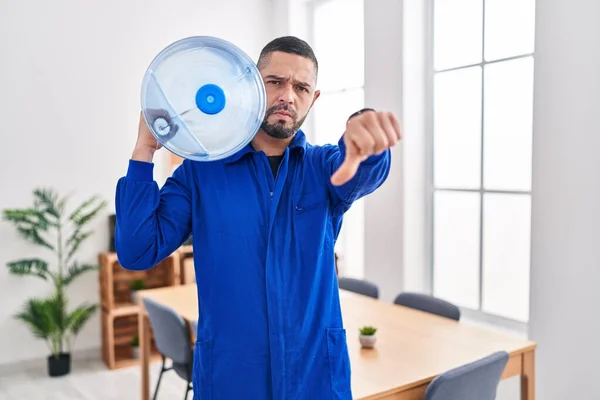 Hispanic service man holding a gallon bottle of water for delivery with angry face, negative sign showing dislike with thumbs down, rejection concept