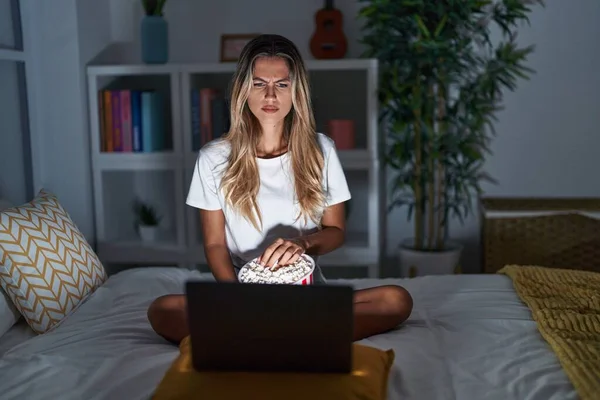 年轻的金发女人坐在床上 坐在家里看一部来自笔记本电脑的电影 充满怀疑和紧张 因为问题而皱着眉头 消极的人 — 图库照片