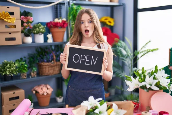 Caucasian woman working at florist holding open sign in shock face, looking skeptical and sarcastic, surprised with open mouth