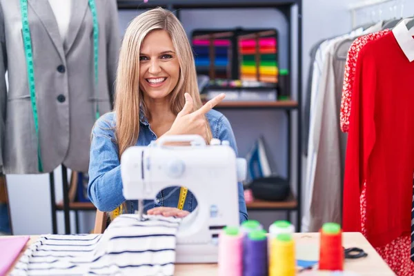 Blonde woman dressmaker designer using sew machine cheerful with a smile on face pointing with hand and finger up to the side with happy and natural expression