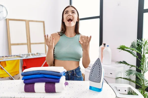 洗濯室で若いヒスパニック系の女性が服をアイロンをかけ 狂った叫びと積極的な表現と腕を上げて叫んでいた フラストレーションコンセプト — ストック写真