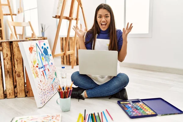 年轻的黑发女子坐在艺术工作室的地板上 手持笔记本电脑 开心地微笑着庆祝胜利 举起双手庆祝胜利 — 图库照片