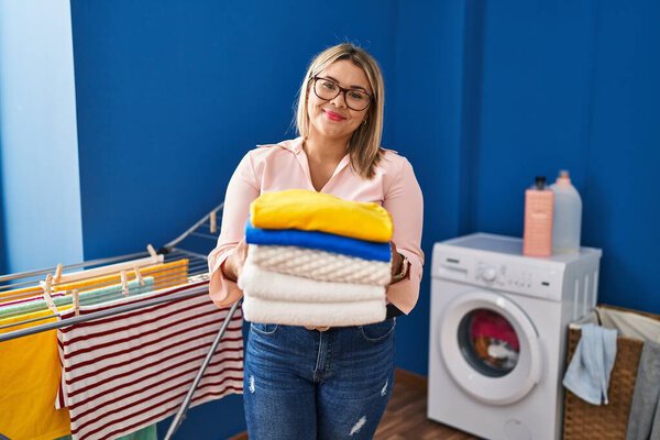 Young hispanic woman holding folded laundry after ironing relaxed with serious expression on face. simple and natural looking at the camera. 