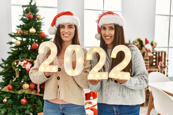 Two women celebrate new year holding 2022 lights standing by christmas tree at home