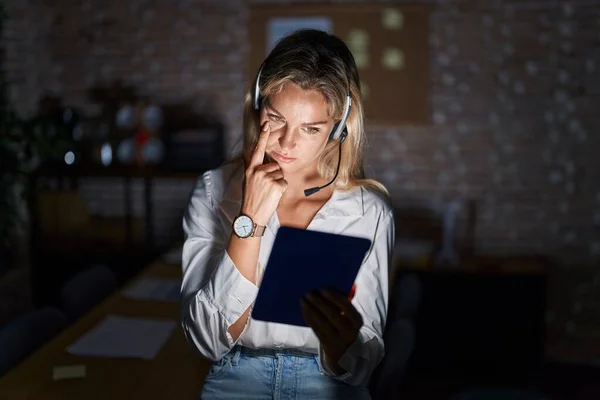 Young blonde woman working at the office at night pointing to the eye watching you gesture, suspicious expression