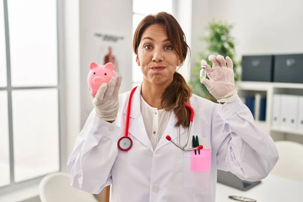 Hispanic Doctor Woman Holding Piggy Bank Puffing Cheeks Funny Face — Stock Photo, Image