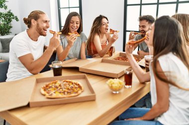 Gülümseyen bir grup genç evdeki masada oturmuş İtalyan pizzası yiyor.