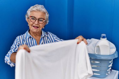 Çamaşır odasında beyaz t-shirt tutan yaşlı, gri saçlı bir kadın gülümsüyor.