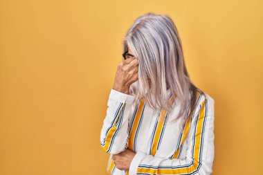 Sarı arka planda duran gri saçlı, gözlüklü, burnu ve gözleri ovuşturmaktan yorgun, yorgun ve başı ağrıyan orta yaşlı bir kadın. Stres ve hüsran kavramı. 