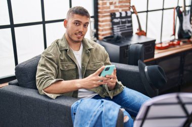 Young hispanic man musician using smartphone at music studio
