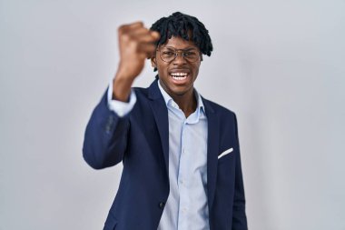 Young african man with dreadlocks wearing business jacket over white background angry and mad raising fist frustrated and furious while shouting with anger. rage and aggressive concept. 