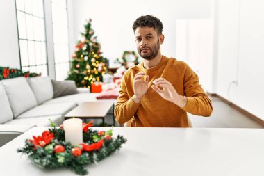 Arap genç adam, yılbaşı ağacının yanında masada oturuyordu. Tiksinti tepkisi yüzünden tiksinmiş ve ürkek bir ifade takınıyordu. Eller havada. 