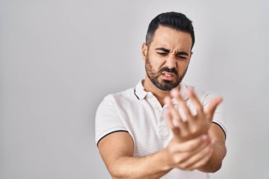 Beyaz arka plan üzerine günlük giysiler giyen, elleri ve parmakları ağrıyan, eklem iltihabı olan Latin kökenli genç bir adam. 