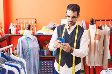 Young hispanic man tailor using smartphone at atelier