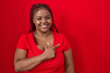 African american woman with braided hair standing over red background cheerful with a smile of face pointing with hand and finger up to the side with happy and natural expression on face 