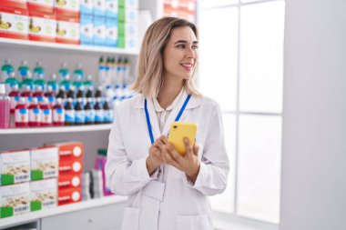 Young woman pharmacist smiling confident using smartphone at pharmacy