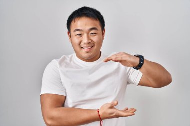 Young chinese man standing over white background gesturing with hands showing big and large size sign, measure symbol. smiling looking at the camera. measuring concept. 