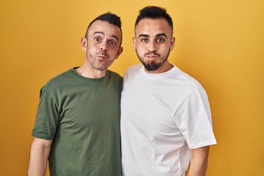 Homosexual couple standing over yellow background puffing cheeks with funny face. mouth inflated with air, crazy expression. 