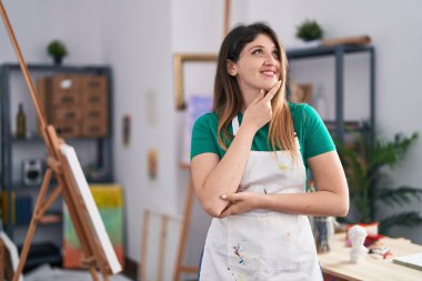 Sanat stüdyosundaki genç esmer kadın kameraya kollarını kavuşturmuş ve çenesinde elle gülümsüyor. Olumlu düşünmek. 